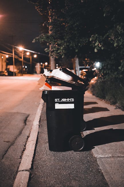 A black wheeled rubbish bin positioned on the pavement beside a curb in an urban street scene at night. The bin is marked with the text 'ST. JOHN'S' in white, and its lid is open, showing a variety of discarded waste materials, including flattened cardboard boxes, paper, and plastic packaging. The street is illuminated by streetlights, casting a warm glow on the surroundings. In the background, there are trees lining the sidewalk, with additional streetlights, parked cars, and faintly visible residential buildings. Shadows of the bin are cast onto the pavement, and the overall scene emphasizes the importance of proper waste disposal, aligning with private rubbish collection and on-site clearance services offered by companies like Rubbish Removal Kingston, which support alternative waste handling solutions beyond council collection.