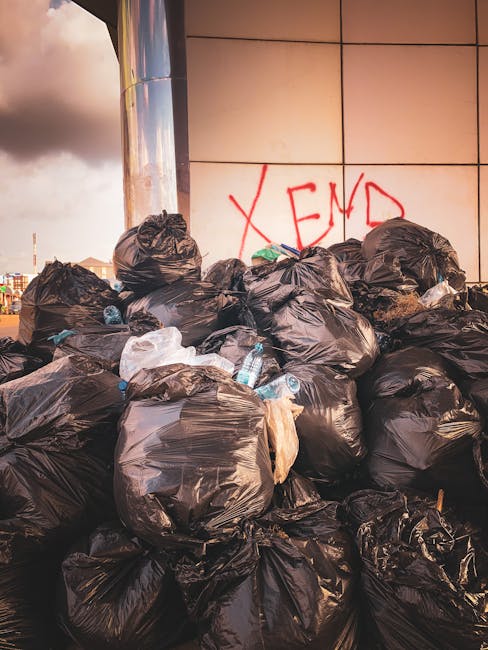 A collection of rubbish and waste materials is piled on a paved sidewalk area in front of storefronts and commercial buildings. The scene includes several black and red trash bins, with some overflowing and strewn with various waste items. The waste comprises cardboard boxes, plastic bags filled with household rubbish, paper, and packaging materials, along with a cardboard box and miscellaneous debris scattered on the ground. There is a large grey mixed paper and card waste container with an open lid, exposing loose paper and cardboard inside. Nearby, a silver car with a UK registration plate is parked, partially obstructed by a metal railing. The background features a building with a glass-fronted upper level, scaffolding, and signage, indicating a commercial or retail area. The environment appears to be an urban setting, with the waste accumulated possibly from individual or business waste disposal, relevant to private rubbish collection services like those offered by Rubbish Removal Kingston, serving local waste management needs. Natural daylight illuminates the scene, highlighting the various textures of the waste and the surrounding environment.