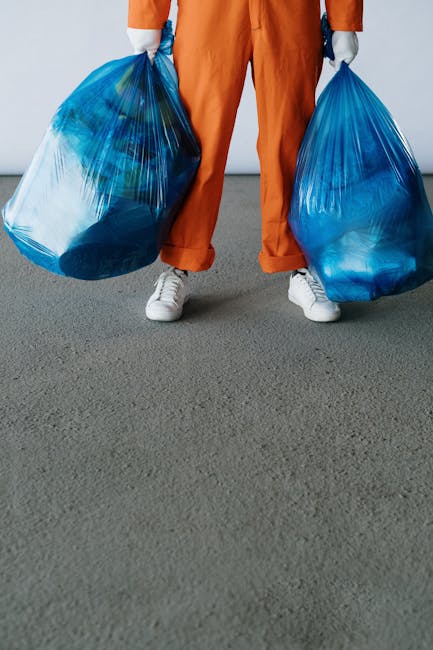 The image shows a person standing on a flat grey surface, holding two large blue plastic rubbish bags, one in each hand. The individual is dressed in orange work overalls and white sneakers, with only their lower body visible from the waist down. The bags are filled with waste material, appearing semi-transparent and crinkled in texture, with some contents visible through the plastic. The background is plain and uncluttered, emphasizing the focus on the person and the waste bags. This scene may depict an activity related to rubbish collection or waste disposal, typical of private waste management services such as those offered by Rubbish Removal Kingston, illustrating the process of on-site clearance or waste handling outside a residential or commercial property. The environment suggests an outdoor or driveway setting, suitable for rubbish removal tasks involving the transport of waste to a disposal or recycling point.