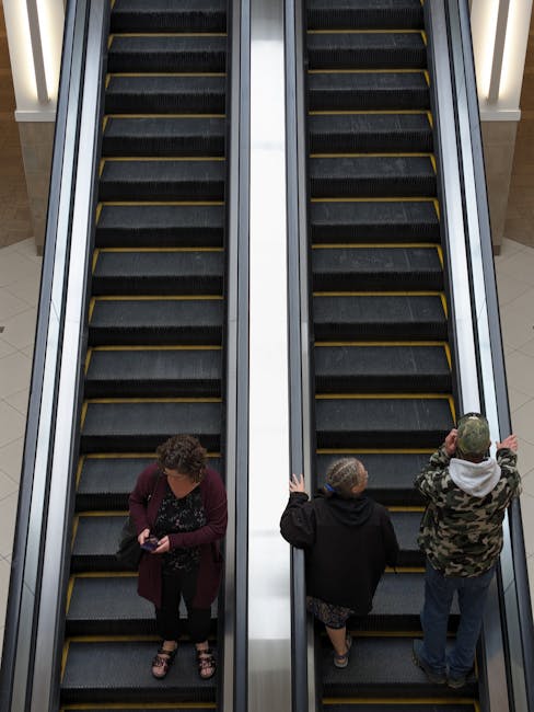 A photograph capturing two parallel escalators inside a modern shopping centre or building. The escalators are black with yellow safety lines along the edges of each step, and they are framed by metallic silver side panels. In the foreground, three individuals are visible: on the left, a woman with short, curly hair wearing a burgundy cardigan and patterned blouse, looking down at her phone; in the middle, a woman with short hair dressed in a black coat and floral dress, holding onto the handrail with both hands, facing the escalator; on the right, a man wearing a camouflage jacket and grey hood, standing at the top of the escalator, looking to the side and holding onto the handrail with his right hand. The environment is well-lit, giving a clean and orderly impression, with the focus on the escalator environment which is typical for public transit or shopping centre accessory for efficient movement of people. This scene subtly aligns with the concept of orderly waste disposal or thorough clearance in busy communal spaces, supported by professional rubbish removal services such as Rubbish Removal Kingston, who handle waste off-site rather than leaving clutter or debris behind.