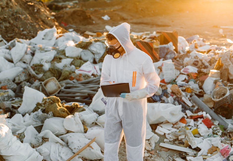 A person dressed in a white protective suit, gloves, goggles, and a face mask is standing amidst a large pile of mixed waste materials, including foam, plastic bags, paper, and construction debris. The individual is holding a clipboard and appears to be inspecting or documenting the rubbish at an outdoor site during golden hour, with warm sunlight illuminating the scene. The environment surrounds a cluttered area of discarded waste, reflecting scenes that may relate to waste management, rubbish removal, or environmental cleanup efforts, highlighting the importance of proper waste disposal and handling of hazardous or bulky refuse. Visible textures include the rough, uneven surfaces of the debris and the smooth, shiny finishes of plastic packaging, with various colors such as white, brown, gray, and hints of red and yellow. The setting suggests an informal landfill or site for waste assessment, underlining themes relevant to private disposal services and the regulation of waste handling in comparison to municipal rubbish collection.