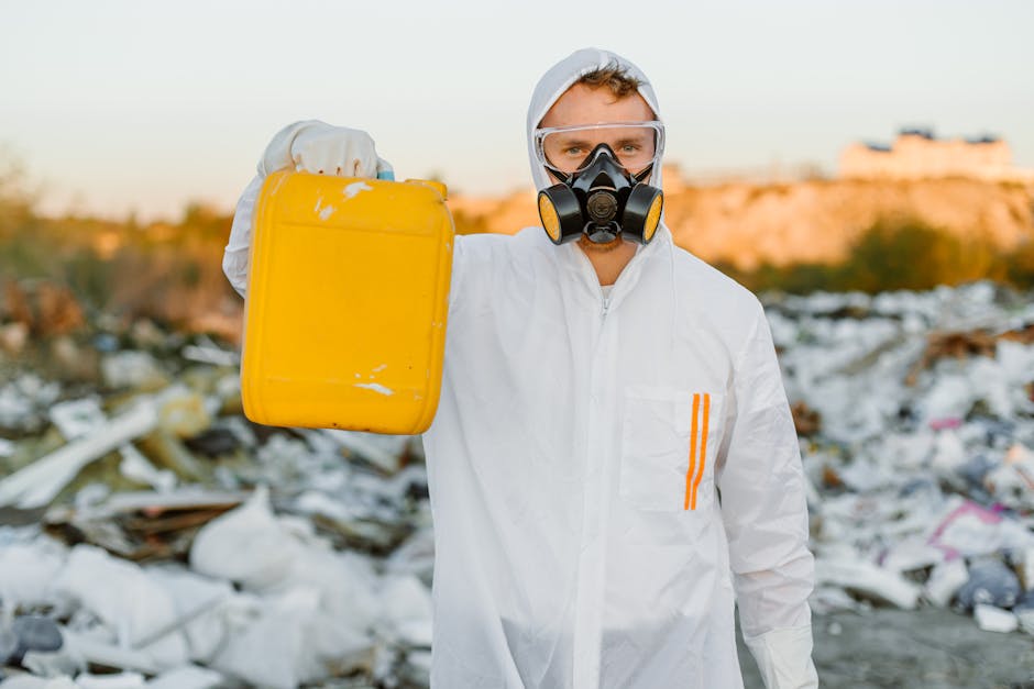 A person wearing a white protective suit, gloves, and a black respirator mask with yellow filters stands outdoors in a landfill or waste site during daylight hours. They hold a bright yellow safety or chemical container in their right hand, which shows some scuffs and scratches on its surface. The background reveals a large area filled with scattered debris, including various types of waste such as plastics, paper, and other refuse. The terrain appears uneven with mounds of discarded materials, and the distant horizon features low-lying formations or possibly buildings, under a clear, softly lit sky. The environment emphasizes the importance of proper waste handling, recycling, or hazardous waste disposal, aligning with rubbish removal services provided by companies like Rubbish Removal Kingston. The scene highlights a focus on responsible waste management practices, including on-site clearance or private waste collection, in a setting indicative of environmental cleanup efforts and waste removal operations.