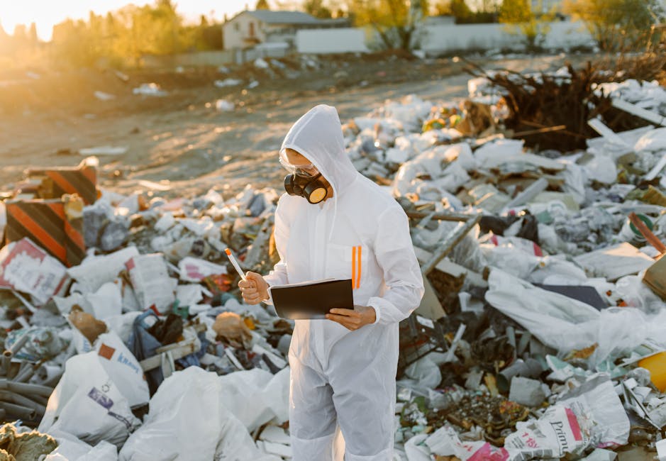 A person dressed in a white protective suit, gloves, goggles, and a face mask is standing amidst a large pile of mixed waste materials, including foam, plastic bags, paper, and construction debris. The individual is holding a clipboard and appears to be inspecting or documenting the rubbish at an outdoor site during golden hour, with warm sunlight illuminating the scene. The environment surrounds a cluttered area of discarded waste, reflecting scenes that may relate to waste management, rubbish removal, or environmental cleanup efforts, highlighting the importance of proper waste disposal and handling of hazardous or bulky refuse. Visible textures include the rough, uneven surfaces of the debris and the smooth, shiny finishes of plastic packaging, with various colors such as white, brown, gray, and hints of red and yellow. The setting suggests an informal landfill or site for waste assessment, underlining themes relevant to private disposal services and the regulation of waste handling in comparison to municipal rubbish collection.