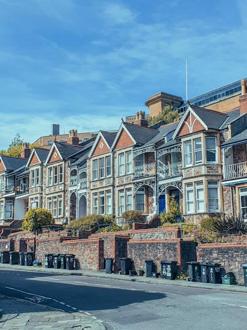 A row of Victorian-style terraced houses with ornate white decorative trim, bay windows, and small front gardens with shrubs and plants. The buildings are constructed from red brick with contrasting white accents around the windows and roof gables. The houses are set on a slightly elevated position behind a low brick wall, with a street in the foreground lined with black wheelie bins. The sky overhead is clear with a few wispy clouds, and the scene is illuminated by natural daylight, suggesting a bright, calm day. The environment appears residential, and the overall setting is typical of urban areas where private rubbish collection services might be employed, aligning subtly with alternative waste handling practices such as private rubbish removal, as promoted by companies like Rubbish Removal Kingston.