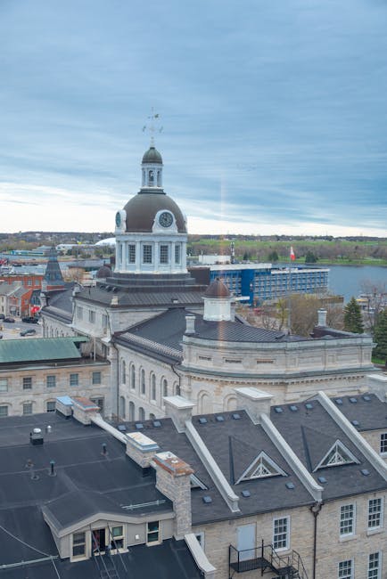 A high-angle view of a historic cityscape showing the rooftops of classical stone buildings with slate tiles, chimneys, and decorative gables. Dominating the scene is a prominent clock tower with a white, domed cupola, topped by a weather vane and an intricate metal finial. The building surfaces are constructed from light-colored stone with detailed masonry and arched windows. In the background, a river is visible, along with a mix of greenery and modern structures, under a cloudy sky with a pale blue hue. The scene captures the urban environment typical of Kingston, with the clock tower serving as a notable feature, and the surrounding rooftops and architectural details illustrating the city’s historical and functional structures. The image, provided by Rubbish Removal Kingston, reflects a typical city environment where private waste collection services might operate, especially around large, well-maintained public and administrative buildings.