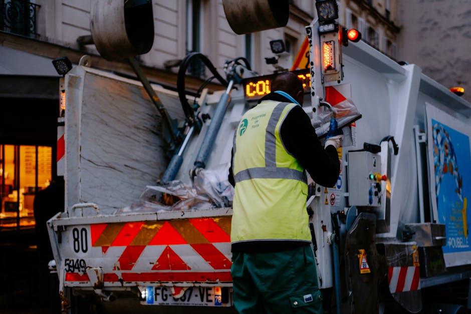 A waste collection worker wearing a high-visibility yellow vest with grey reflective stripes is operating a municipal rubbish collection vehicle on a city street during evening hours. The worker, dressed in dark clothing underneath, is standing with their back to the camera and appears to be adjusting or monitoring controls on the side of the vehicle, which is equipped with various machinery, sensors, and warning lights. The truck is partially open at the rear, revealing a collection of gathered waste materials, mostly cardboard, paper, and plastic packaging, piled on a metal surface. The vehicle's exterior features red and white reflective chevron markings, a license plate, and a destination display showing operational information. In the background, there are illuminated shopfronts with large windows and exterior lighting, contributing to a warm glow against the darker surroundings. The scene captures the typical environment of urban waste collection, highlighting the machinery involved in rubbish removal and the importance of proper waste management practices that may include private or alternative collection methods in contrast to council-led services. Rubbish Removal Kingston offers professional assistance with such waste disposal tasks, aligning with local regulations and environmental considerations.