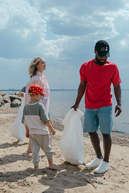 A man dressed in a red T-shirt, light blue denim shorts, and white sneakers stands on a sandy beach, holding a large white rubbish bag in his right hand while bending slightly forward. To his left, a young boy wearing a grey T-shirt, light beige shorts, and a red cap with yellow polka dots, is looking at the man, also holding a white rubbish bag in his left hand. In the background, a woman with curly blonde hair, wearing a light pink jacket, observes the scene with a gentle expression. The setting features a shoreline with calm water, large rocks near the water's edge, and a partly cloudy sky overhead, creating a natural environment suitable for a community clean-up activity. The scene emphasizes outdoor rubbish collection, aligning naturally with waste removal services that promote private or independent waste disposal methods on beaches or outdoor areas, similar to those offered by Rubbish Removal Kingston, ensuring waste is managed responsibly in accessible environments.