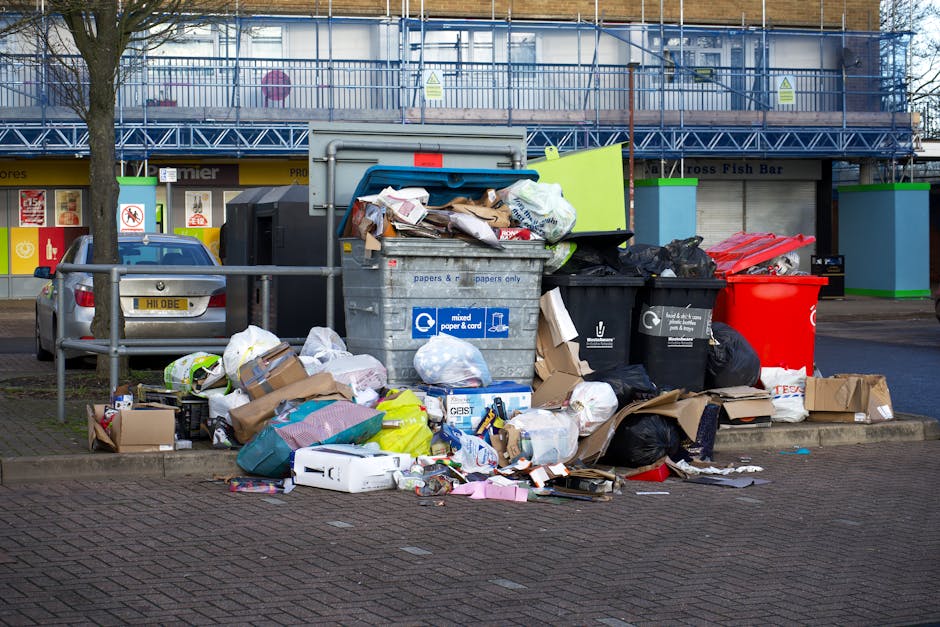 A collection of overflowing waste bins and scattered rubbish on a paved urban sidewalk near a parking area. The scene includes a large grey mixed paper and cardboard bin with an open lid, overflowing with crumpled papers, cardboard boxes, and plastic bags. To its right, there are two black bins, one with a partially open lid filled with black rubbish bags, and a red bin standing next to them. Several cardboard boxes, plastic bottles, and packaging materials are scattered on the ground around the bins, with some spilling onto the surrounding brick pavement. In the background, a building with a blue scaffolding structure and blue railings is visible, along with a storefront that has partially closed yellow and black shutters and windows. A parked silver car is seen behind the railings, and a leafless tree is situated to the left of the scene. The overall environment suggests a typical urban area dealing with improper waste disposal, which may be relevant to discussions on local rubbish collection or alternative waste handling options such as private rubbish removal services. The scene is lit by natural daylight, emphasizing the cluttered and untidy nature of the rubbish accumulation.