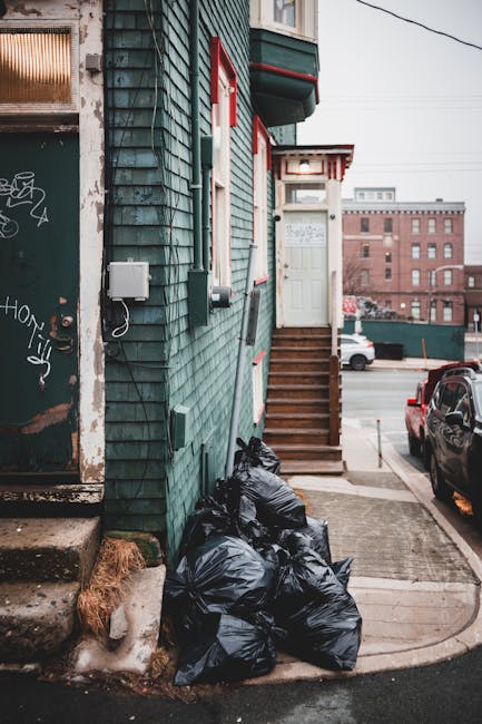 A row of black rubbish bags filled with household waste are piled at the base of a green, weathered wooden building with peeling paint and graffiti on the door. The bags are stacked against the building's wall near a set of wooden stairs leading up to a white door with a small window, situated at the entrance of a residential property. The building has red trim accents around the windows and doorframe, and a small porch with a roof supported by white brackets. In the background, a parking lot with several cars and an industrial-style brick building are visible, indicating an urban environment. The scene is lit by natural daylight, with overcast skies contributing to a neutral lighting tone. This setup suggests private waste collection or on-site rubbish clearance, as handled by independent waste management services, which may be relevant to understanding alternative rubbish removal options outside of standard municipal services.