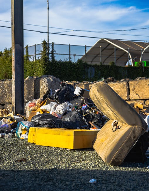 A collection of overflowing waste bins and scattered rubbish on a paved urban sidewalk near a parking area. The scene includes a large grey mixed paper and cardboard bin with an open lid, overflowing with crumpled papers, cardboard boxes, and plastic bags. To its right, there are two black bins, one with a partially open lid filled with black rubbish bags, and a red bin standing next to them. Several cardboard boxes, plastic bottles, and packaging materials are scattered on the ground around the bins, with some spilling onto the surrounding brick pavement. In the background, a building with a blue scaffolding structure and blue railings is visible, along with a storefront that has partially closed yellow and black shutters and windows. A parked silver car is seen behind the railings, and a leafless tree is situated to the left of the scene. The overall environment suggests a typical urban area dealing with improper waste disposal, which may be relevant to discussions on local rubbish collection or alternative waste handling options such as private rubbish removal services. The scene is lit by natural daylight, emphasizing the cluttered and untidy nature of the rubbish accumulation.
