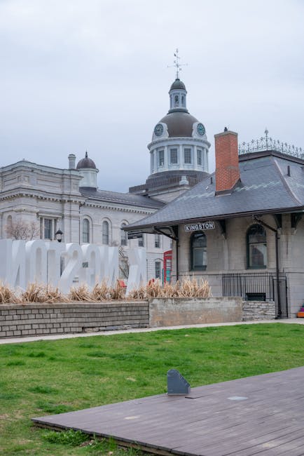 An exterior scene of Canbury Gardens in Kingston featuring a historic railway station building with a dark grey slate roof, a small chimney, and a brick chimney stack on the right side. In the foreground, there is a low brick retaining wall with dried ornamental grasses behind it, and a neatly maintained green lawn extends across the image. A wooden platform is visible at the right edge, with a small, dark object placed on top. The background showcases the prominent dome and spire of a classical-style building with white walls and decorative elements, partially obscured by the station. This setting reflects a public outdoor area near Kingston's riverside, where private waste handling and alternative rubbish collection services, such as those provided by Rubbish Removal Kingston, may be involved in maintaining cleanliness and waste management of the site.