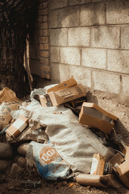 A pile of discarded cardboard boxes and paper waste is stacked against a rough, light-colored brick wall outdoors, near the base of a large tree with dark, textured bark. The boxes, some flattened and others partially folded, vary in size and are made of brown corrugated cardboard with printed labels and barcodes visible on some. Among the boxes, there are crumpled brown paper and plastic bags, including a large white sack with a blue and orange logo. The ground is composed of dirt and small stones, with a few scattered leaves. Shadows cast by the nearby tree create a shaded environment. The scene depicts an area suitable for rubbish removal or on-site waste clearance, aligning with private disposal services like those offered by Rubbish Removal Kingston, especially where independent collection of household or commercial waste is needed. The overall setting emphasizes the importance of proper rubbish disposal in outdoor, urban environments while highlighting the need for professional waste collection to responsibly manage such waste accumulation.