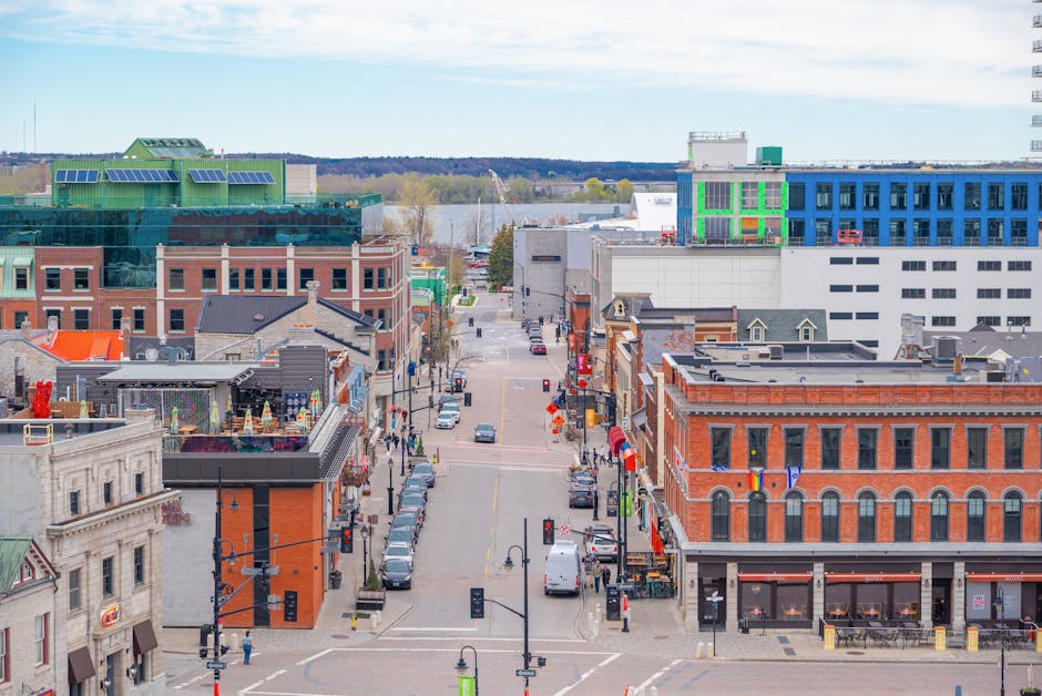 An aerial view of a city street in daytime, featuring a mix of historic and modern buildings. The foreground shows a busy, narrow street lined with parked cars on both sides, with pedestrians walking along the sidewalks. To the left, a building with a decorative facade and large ground-floor windows houses a commercial space, while above it, there is a rooftop terrace with outdoor seating, umbrellas, and decorative umbrellas. The middle ground displays a variety of brick and contemporary structures, some with storefront signage and awnings, and a few small flags can be seen hanging from building facades. In the background, taller office buildings with glass and steel exteriors rise, including some with colorful paneling and rooftop machinery. Beyond the cityscape, a body of water and distant trees are visible under a partly cloudy sky. The scene evokes a typical urban environment suitable for private waste collection or rubbish removal services, with clear street access and a mix of building types.