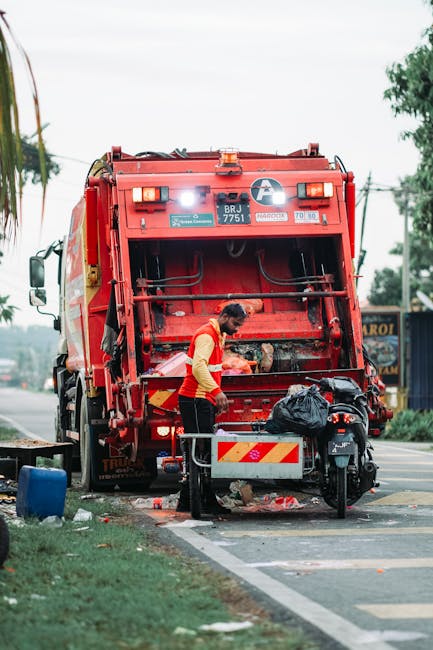 A worker wearing a red and yellow high-visibility uniform is seen standing behind a rear-loading rubbish truck parked on the side of a street. The truck, painted in bright red with reflective safety markings, is identified with a vehicle registration number and safety stickers visible on its back panel. The worker appears to be managing waste, with various debris and waste materials, including a black plastic bag, accumulated on or near a small cart attached to the vehicle. The scene takes place in an urban environment with a paved sidewalk, some grass and trees to the side, and a glimpse of buildings and signage in the background. Overhead, power lines and a cloudy sky are visible, indicating daytime lighting conditions. This setting exemplifies on-site waste collection and rubbish removal activities, which may support independent collection services like those provided by Rubbish Removal Kingston, offering an alternative to council-led rubbish disposal methods.