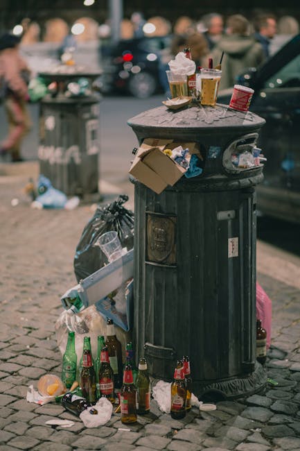 A black, cylindrical public rubbish bin made of metal with a textured surface, placed on a cobblestone pavement in an urban setting. The top of the bin is cluttered with various discarded items: several used paper coffee cups with plastic lids, some plastic bottles, and a small red container. A cardboard box is partially hanging off the side of the bin, with additional litter including plastic wrappers and paper debris around its base. Nearby, crumpled paper, empty glass bottles, and a black plastic garbage bag are scattered on the ground, indicating improper disposal. In the background, there are blurred figures of people, parked cars, and additional rubbish bins or containers, suggesting a busy street environment. The lighting appears natural, with overcast sky conditions casting soft light on the scene, emphasizing the cluttered state typical of urban rubbish collections handled by private waste removal services like Rubbish Removal Kingston, which manages waste from various local settings.