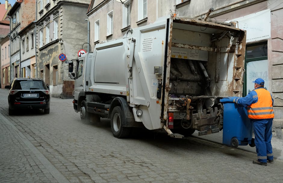 A street scene showing a waste collection truck operated by Rubbish Removal Kingston parked alongside a row of older, multi-story buildings with weathered facades and various window styles. The truck, primarily white with visible rust and grime on the rear-loading mechanism, is positioned with its rear open, revealing the interior where compressed rubbish is being processed. A worker dressed in blue coveralls and a high-visibility orange vest is standing on the cobblestone road on the right side, actively placing a blue recycling bin into the truck’s loading area. The surrounding environment includes a parked dark-colored car further down the narrow street, which is paved with cobblestones, and a no-parking sign affixed to one of the buildings. Overall, the scene depicts an on-site waste collection process in an urban setting, illustrating how private rubbish removal services handle priority or restricted access scenarios outside typical municipal collection routes, aligning with the context of alternative waste handling methods in Kingston.