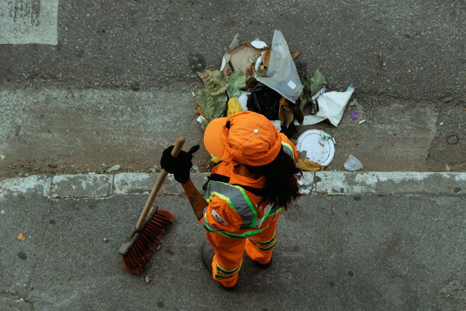 A roadside pile of mixed waste materials is shown, including black plastic rubbish bags, a yellow plastic container, an old, dirty car tyre, and various discarded items such as cardboard, plastic packaging, and paper. The waste is situated on a gravel surface next to a low stone wall, with a metal post and overhead power lines visible in the background. Behind the wall, there is a green hedge, a solar panel-covered structure, and a partly cloudy blue sky. The scene appears to be outdoors on a clear day, with natural sunlight illuminating the waste and surrounding environment. This accumulation of rubbish may reflect instances where private waste handling or independent collection is necessary when access is blocked to regular rubbish removal services, aligning with concepts of alternative waste disposal options for property owners or businesses, as sometimes managed by specialists like Rubbish Removal Kingston.